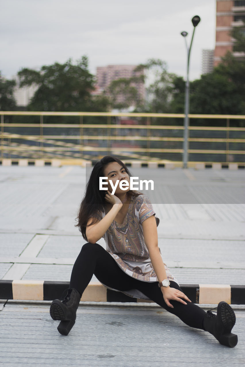 Portrait of young woman with legs apart sitting on sidewalk in city