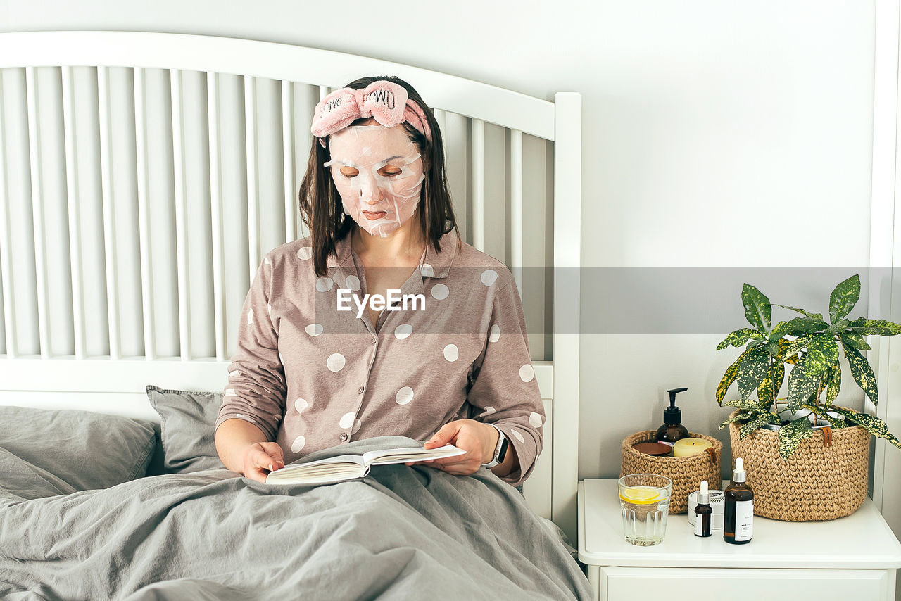 Woman with face mask reading book while while sitting on bed at home