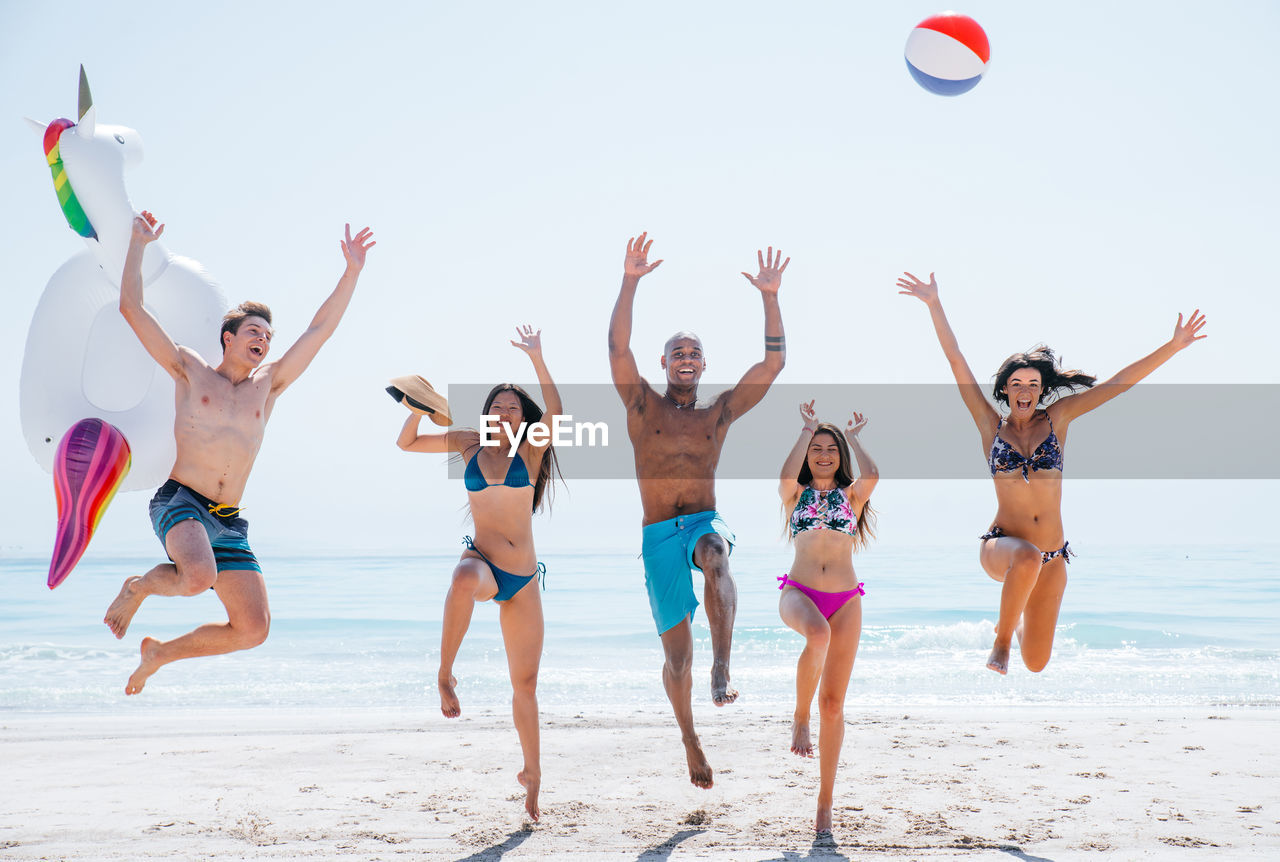 Happy friends jumping on shore of beach against sky