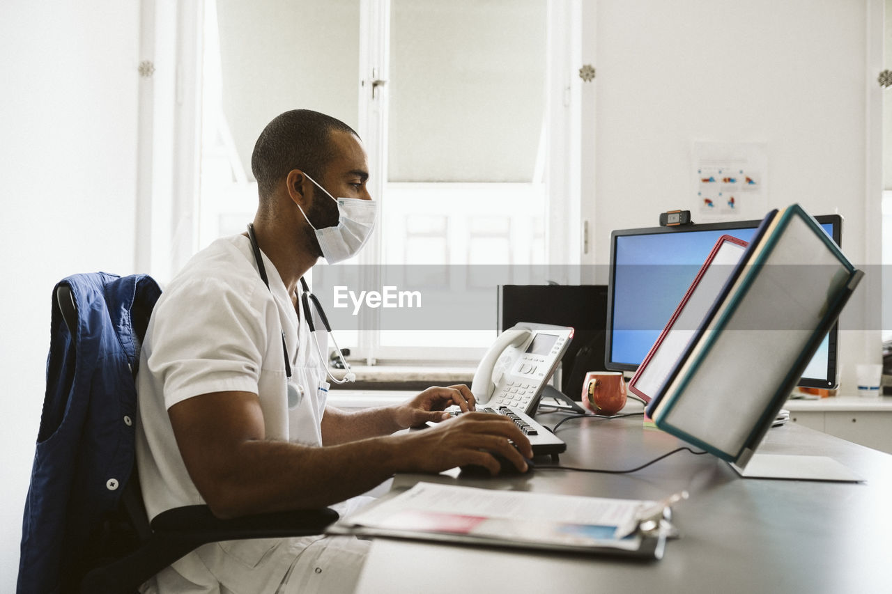 Male doctor using computer while sitting at desk during covid-19