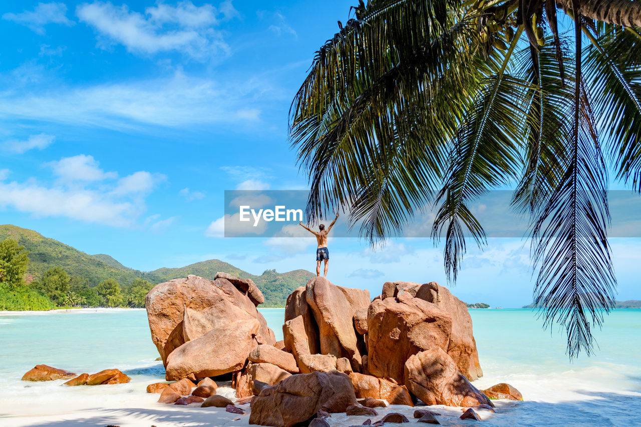 Young man with outstretched hands on rocks over tropical sandy beach.