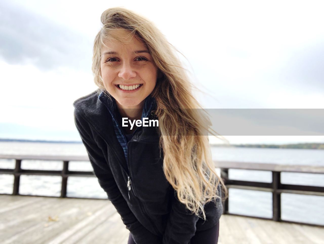 Portrait of young woman smiling while standing on pier over sea against sky