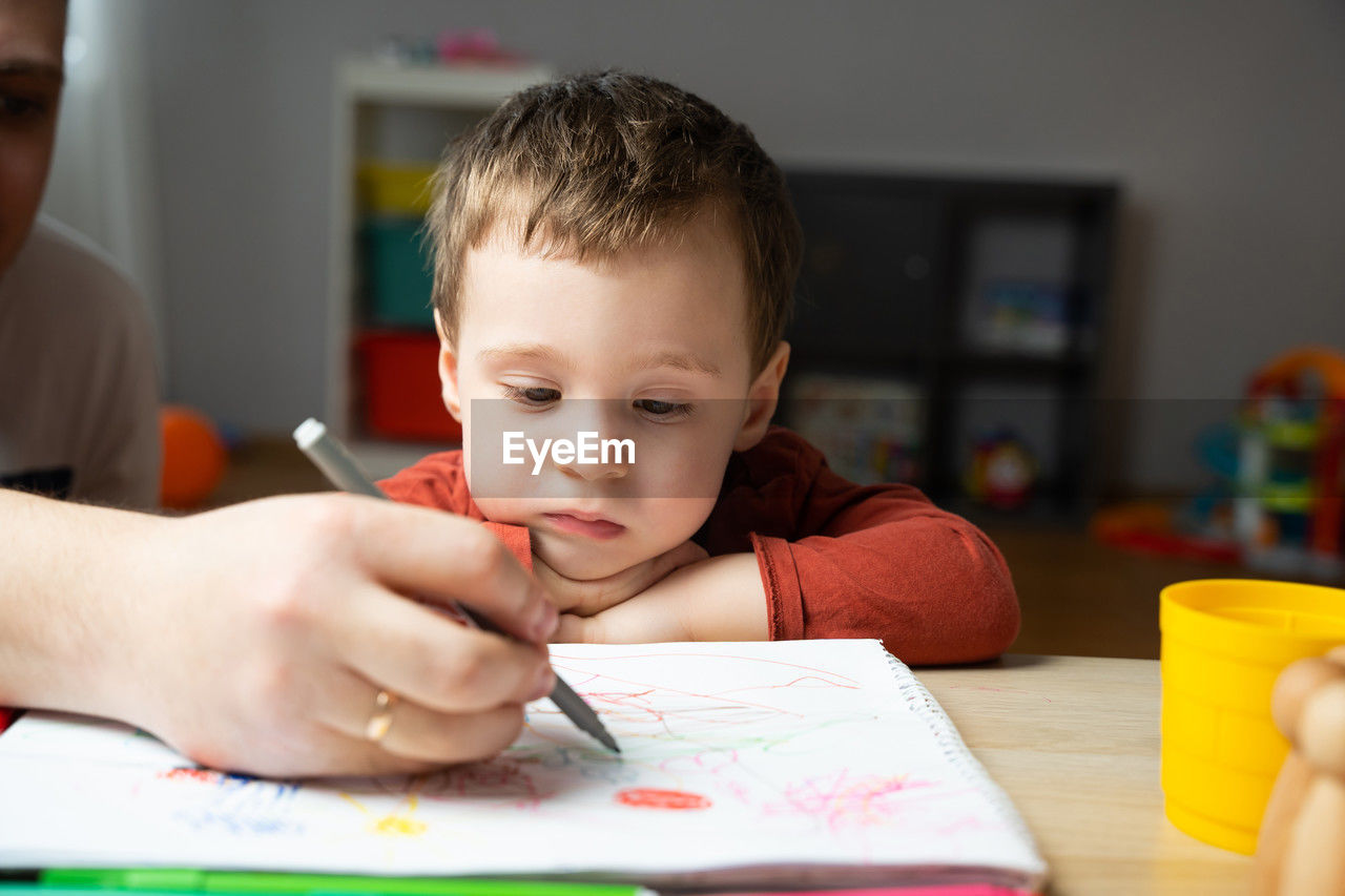 A cute little toddler boy of two years old with dad draws with markers in the album 