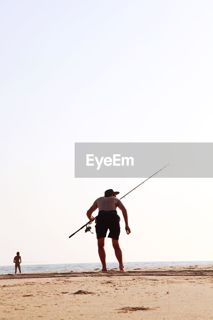 Rear view of shirtless man with fishing rod standing on shore at beach against clear sky