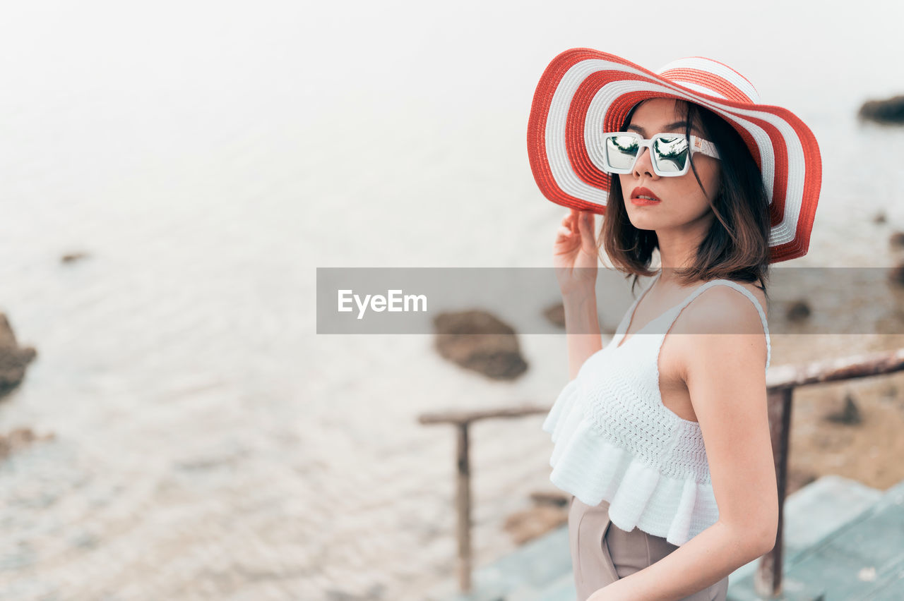 Side view of young woman wearing sunglasses standing against sea