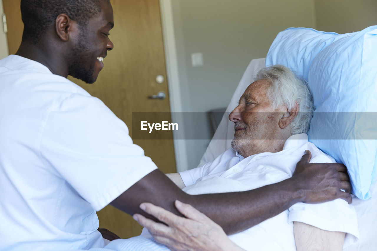 Happy male nurse consoling senior man reclining on bed in hospital ward