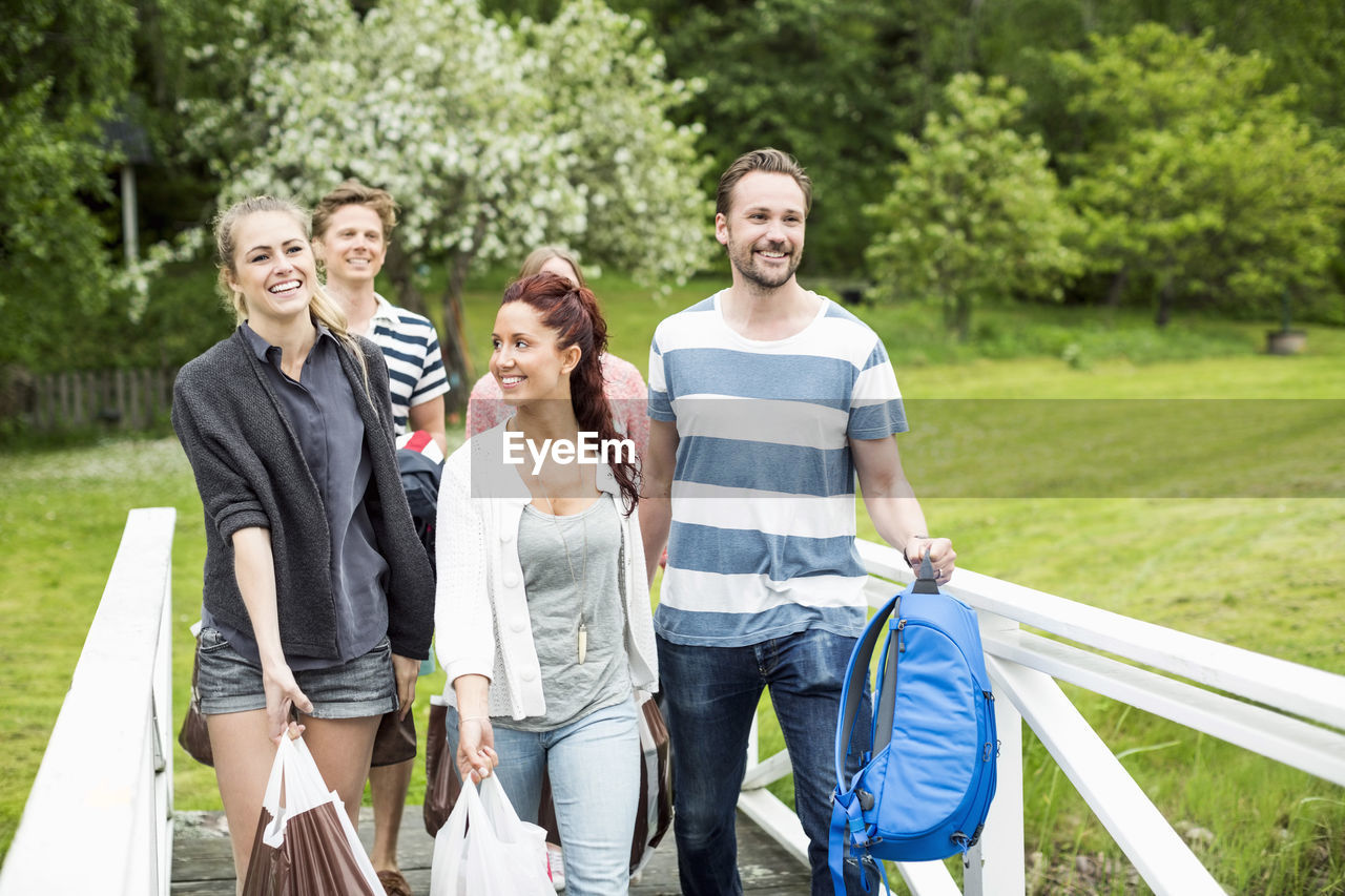 Group of happy friends with luggage walking by railing at park