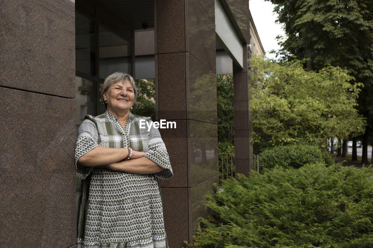 Real cheerful smiling senior woman in yard of modern building. green bushes around. copy space for