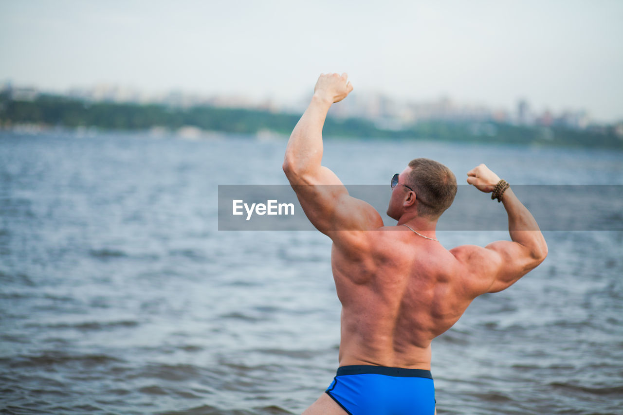 Rear view of shirtless bodybuilder flexing muscles while standing against sea