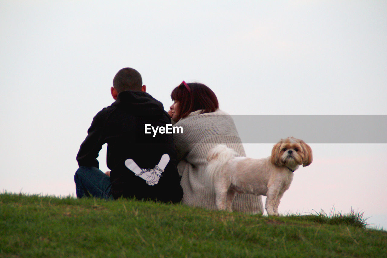 REAR VIEW OF WOMAN WITH DOG SITTING AGAINST SKY