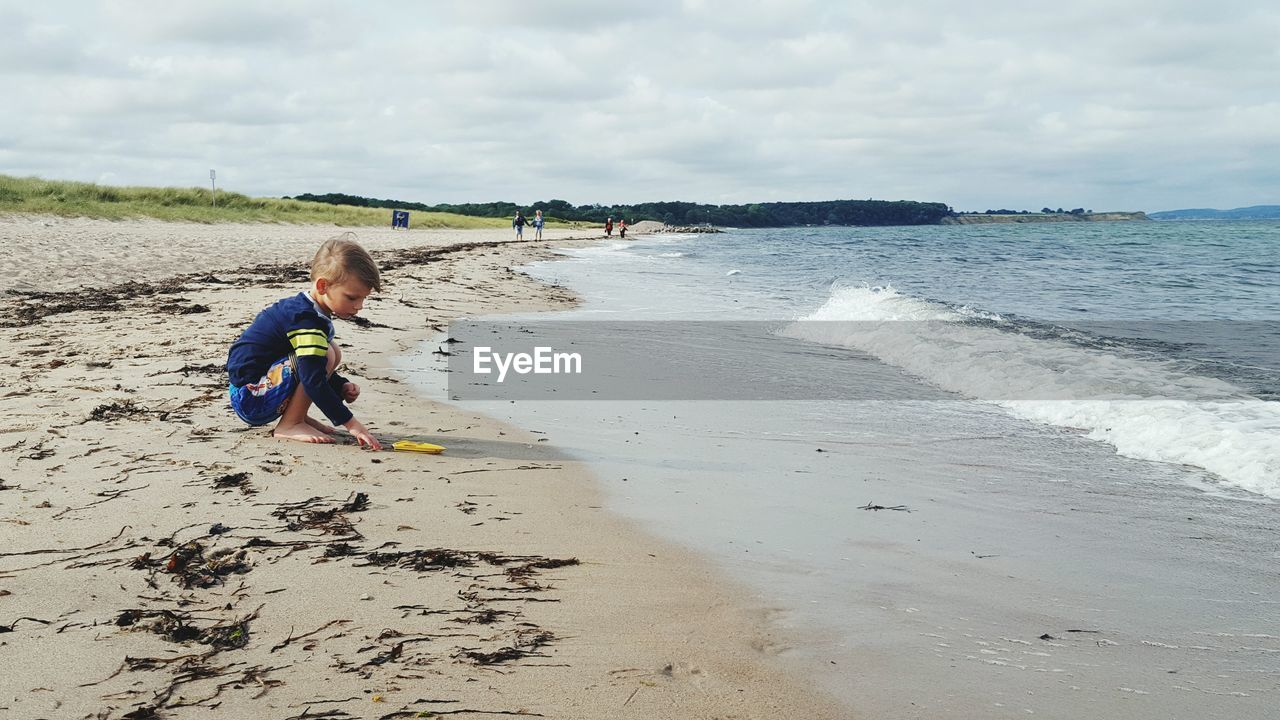 Full length side view of boy touching sand on shore at beach against cloudy sky