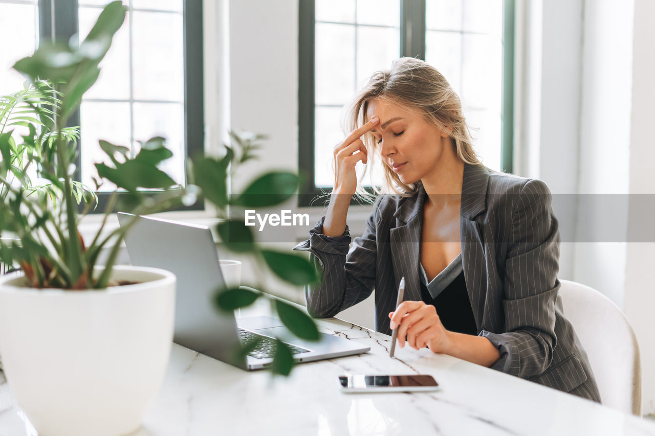 Young tired blonde woman with long hair in stylish grey suit working at laptop in bright office
