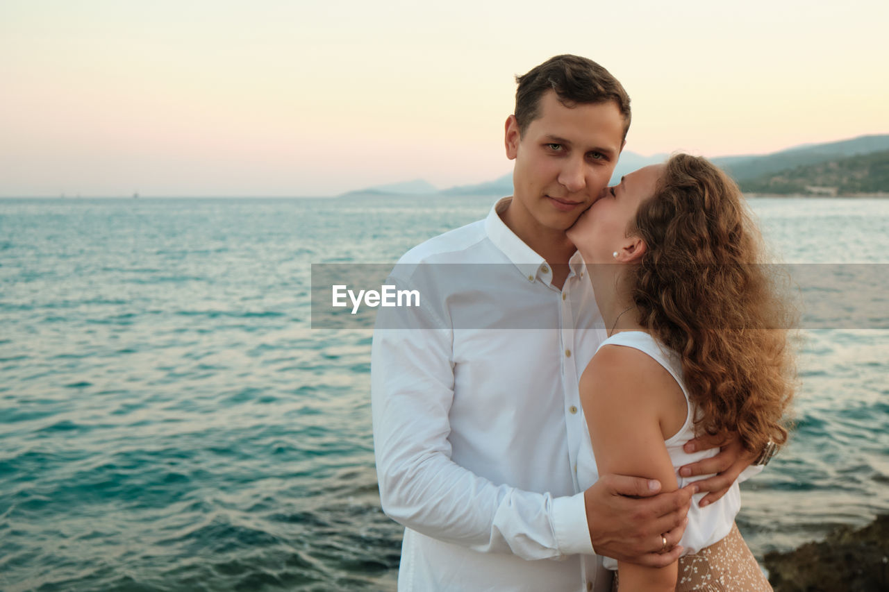 Romantic date on a sea coast. young adult woman kisses her beloved man against seaside sunset