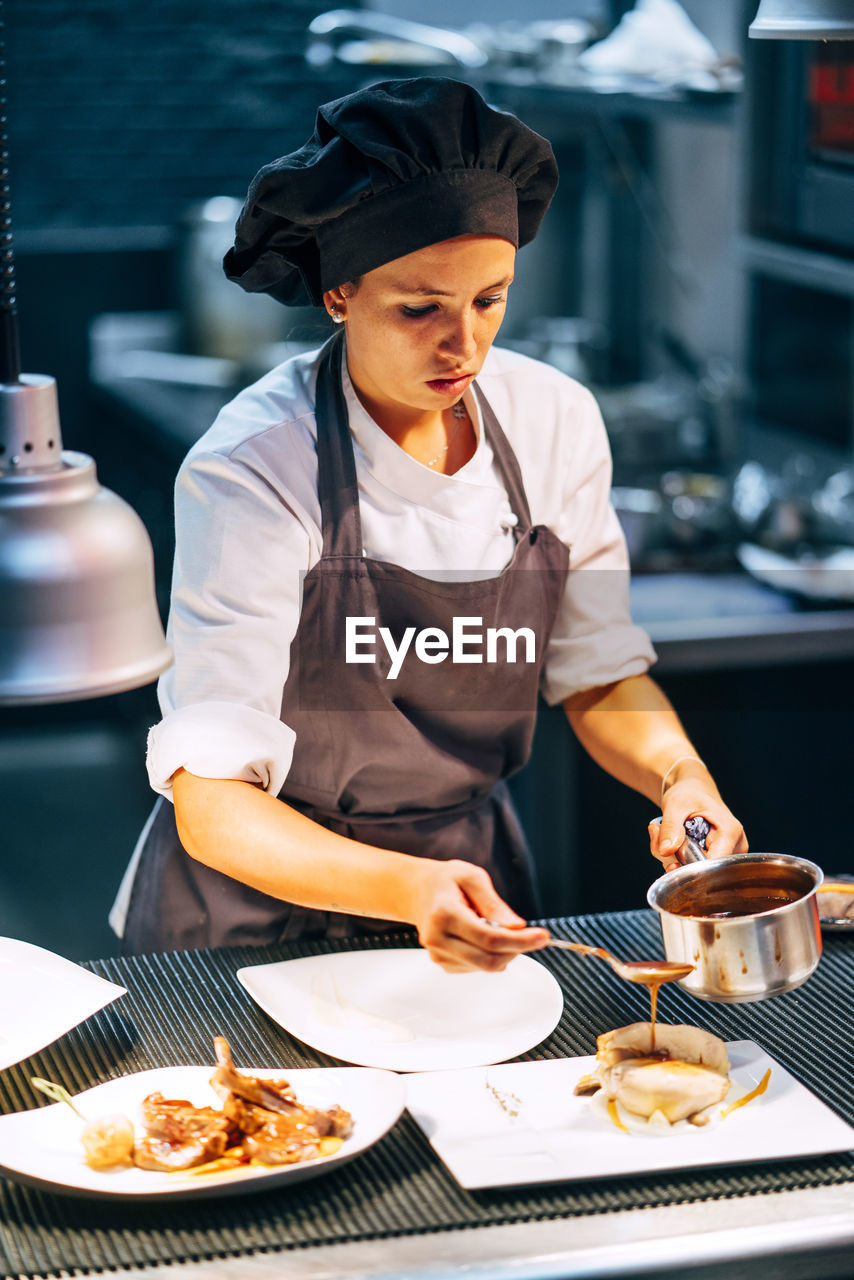 Adult woman in apron and hat preparing and serving dishes on kitchen in modern restaurant