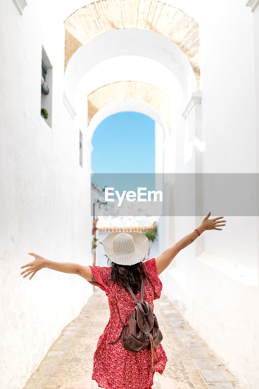 Back view of positive female tourist in straw hat and sunglasses standing with raised arms on narrow street in white village