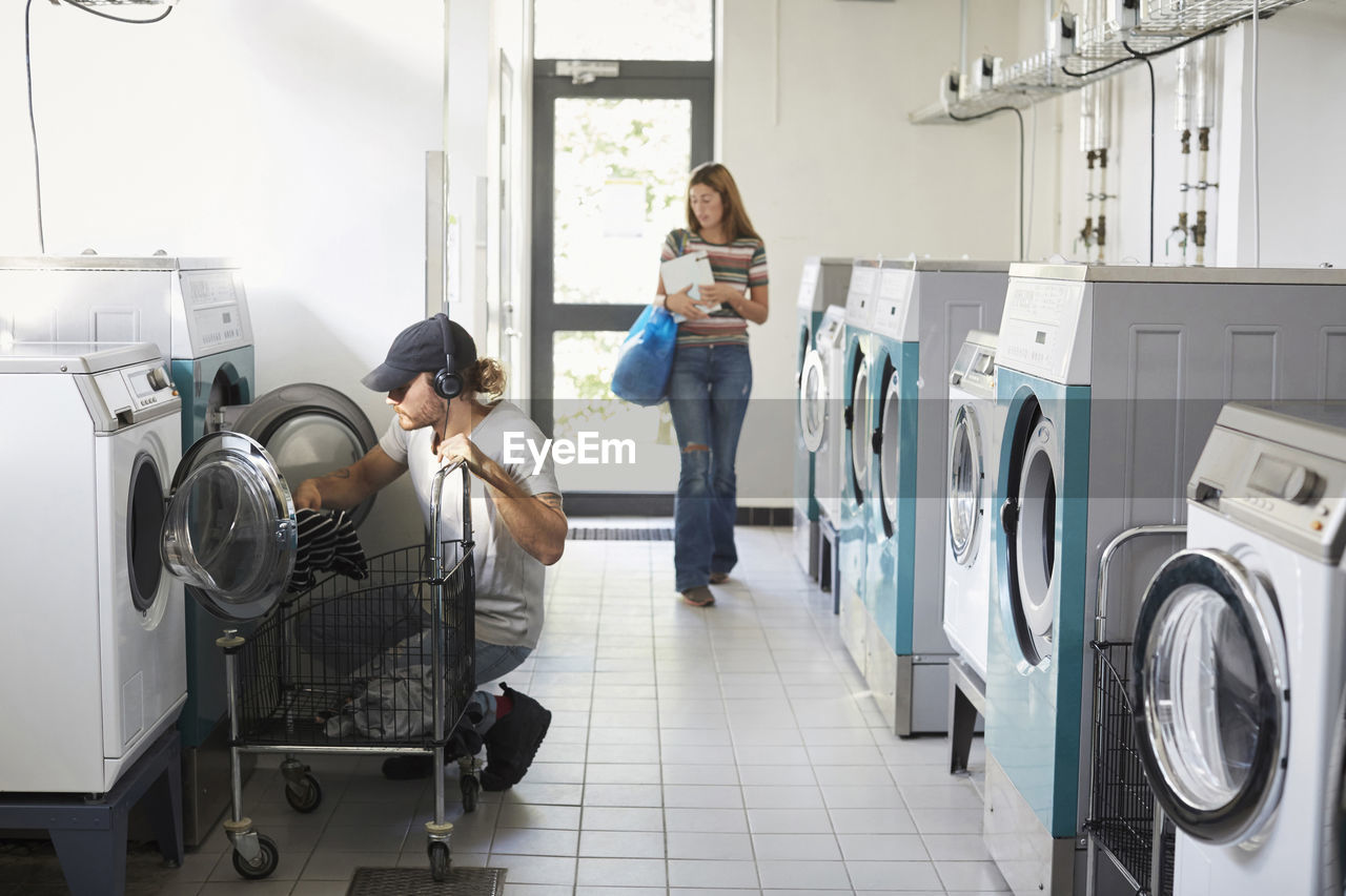 Man using washing machine while woman walking in laundromat
