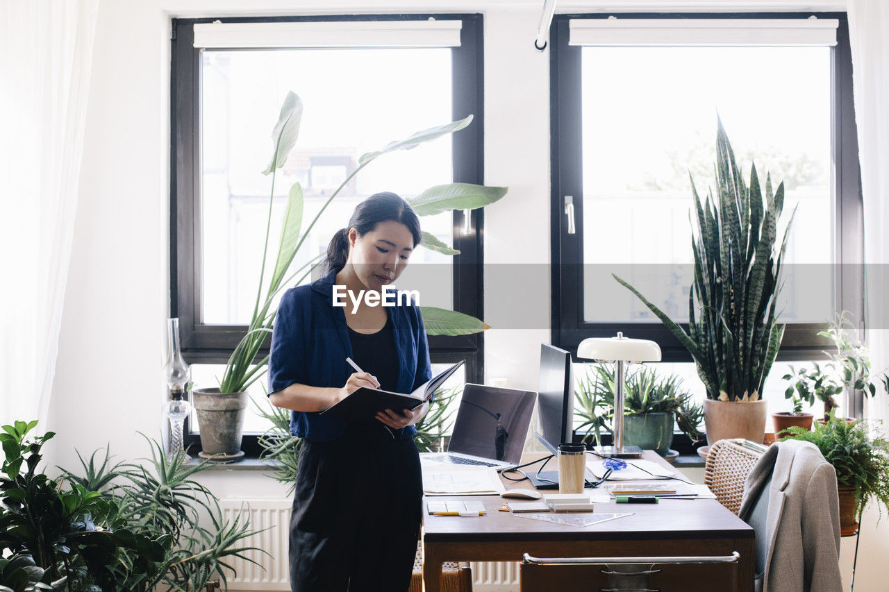 Confident female architect writing in diary while standing by desk in home office