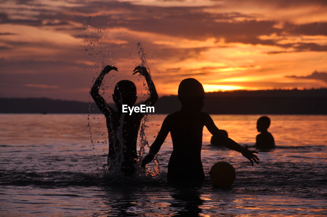 Silhouette children playing in sea against sky during sunset