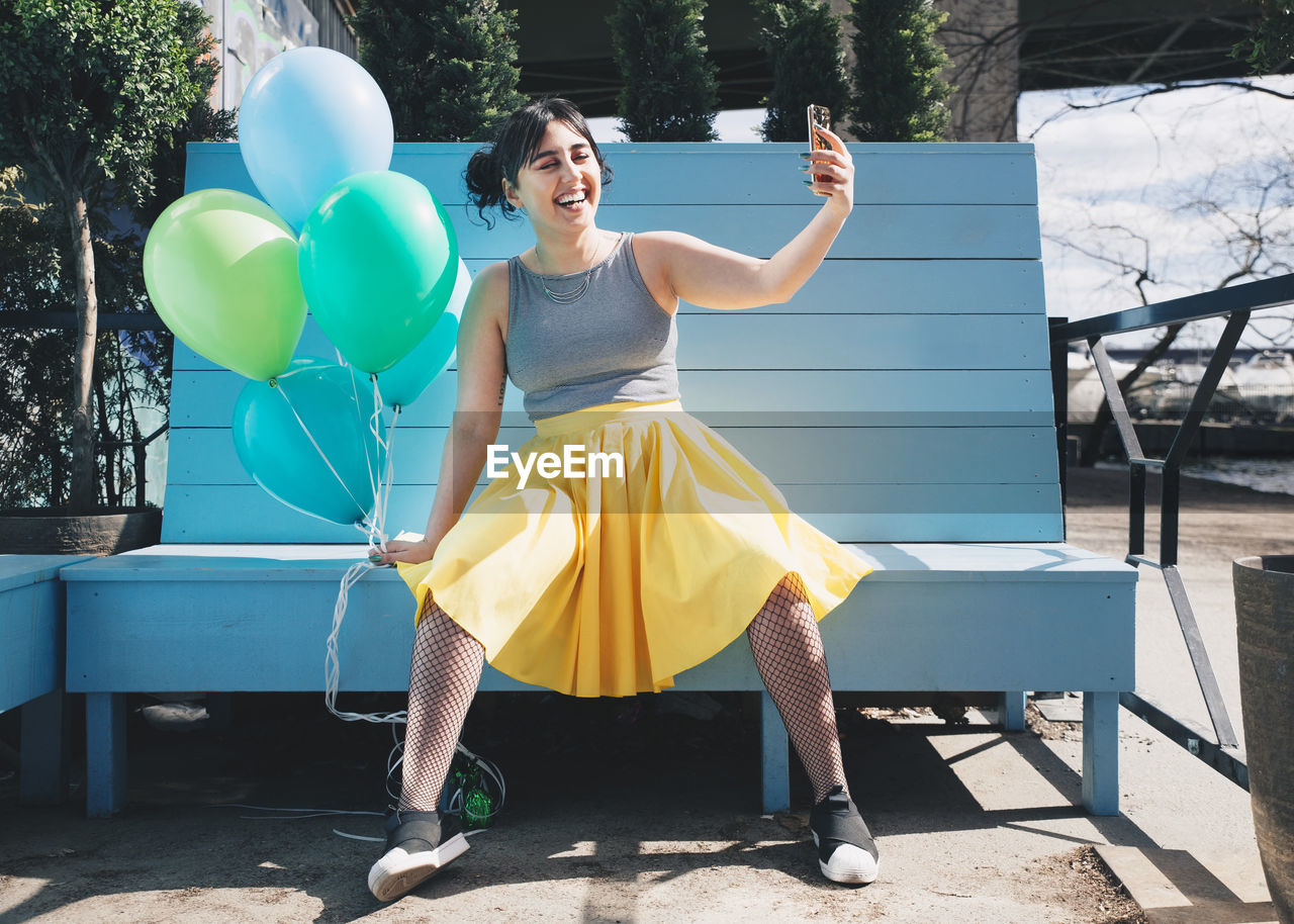 Happy young woman taking selfie with balloons while sitting on bench