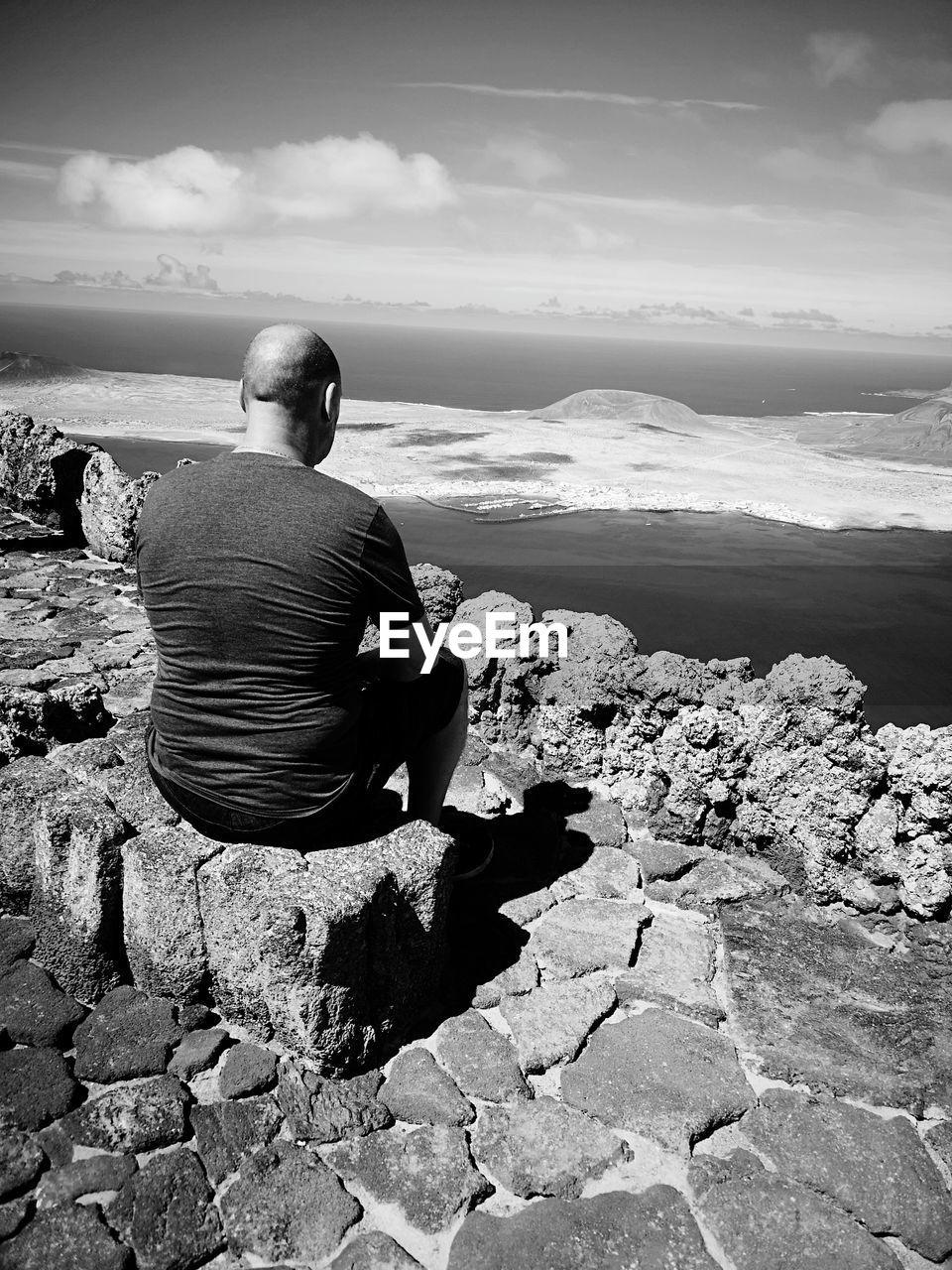 Rear view of man sitting on rock looking at sea against sky