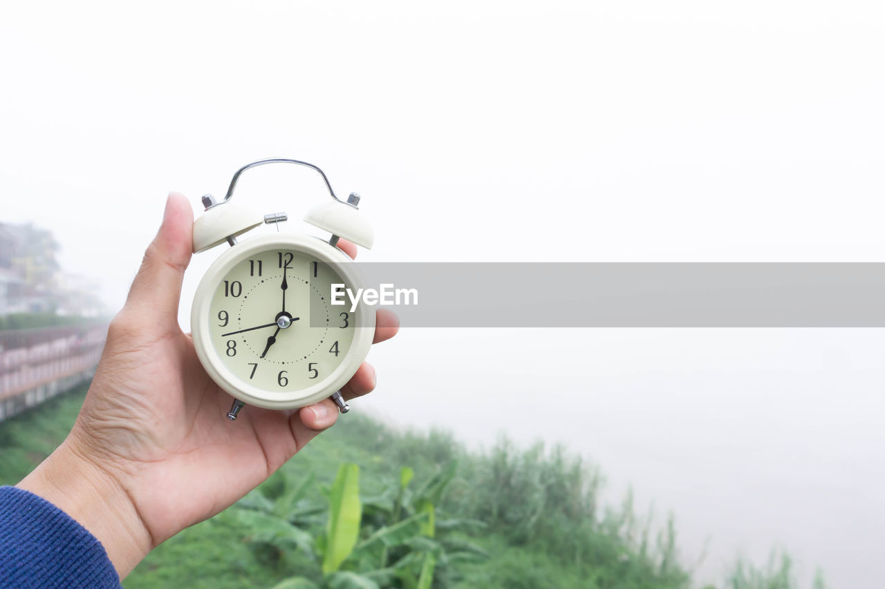 CROPPED IMAGE OF HAND HOLDING CLOCK AGAINST SKY