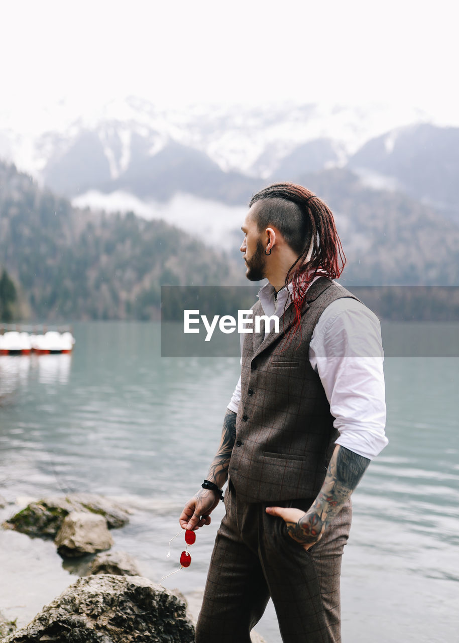 A man a groom a hipster with dreadlocks in a wedding suit stands by sea and mountains in nature