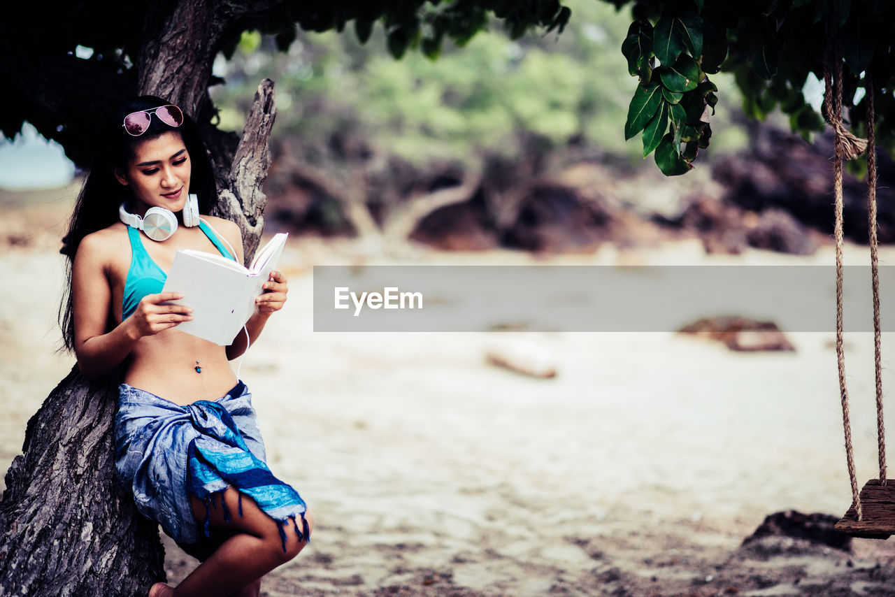 Young woman reading book while leaning on tree at beach