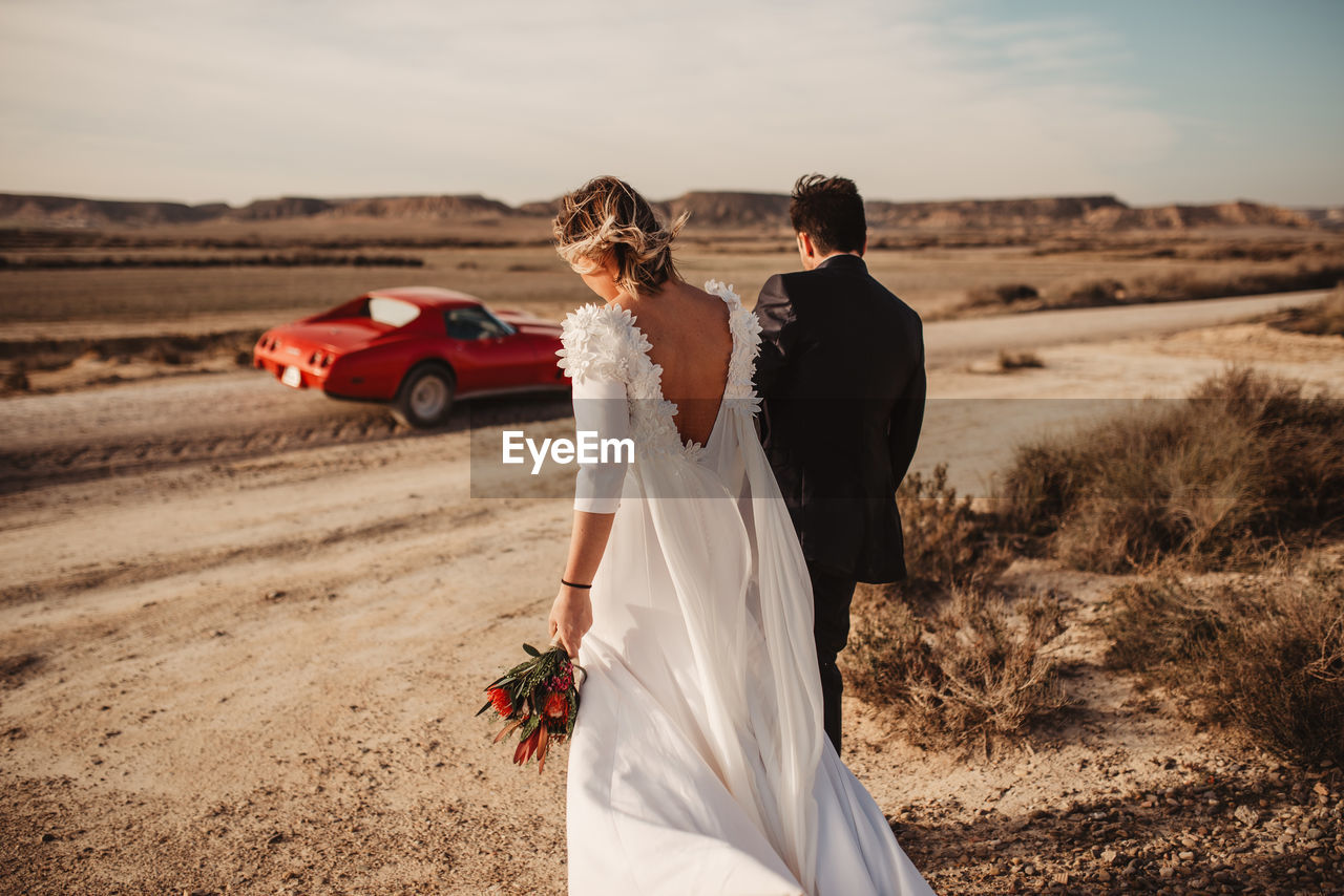 Back view of anonymous bride and groom strolling together towards luxury red car in desert of bardenas reales natural park in navarra, spain