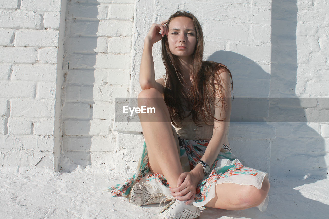 Full length portrait of woman sitting against wall during sunny day