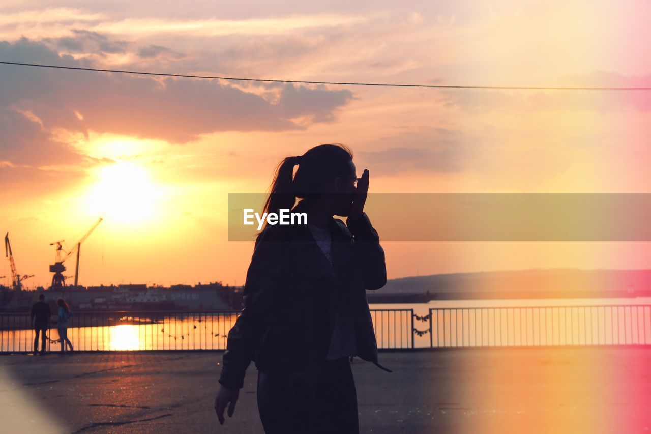 Woman standing on promenade by river against sky during sunset