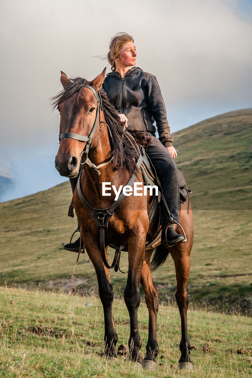 A cowgirl shepherd rider on a horse gallops through a mountain valley on autumn day sky with clouds