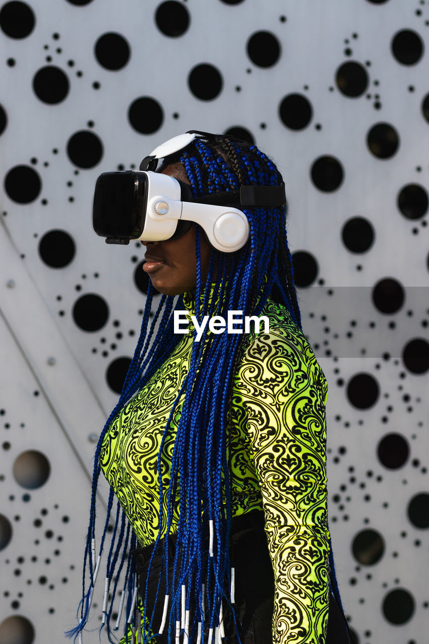 Side view of african american female with blue hair standing in street in vr goggles while experiencing virtual reality