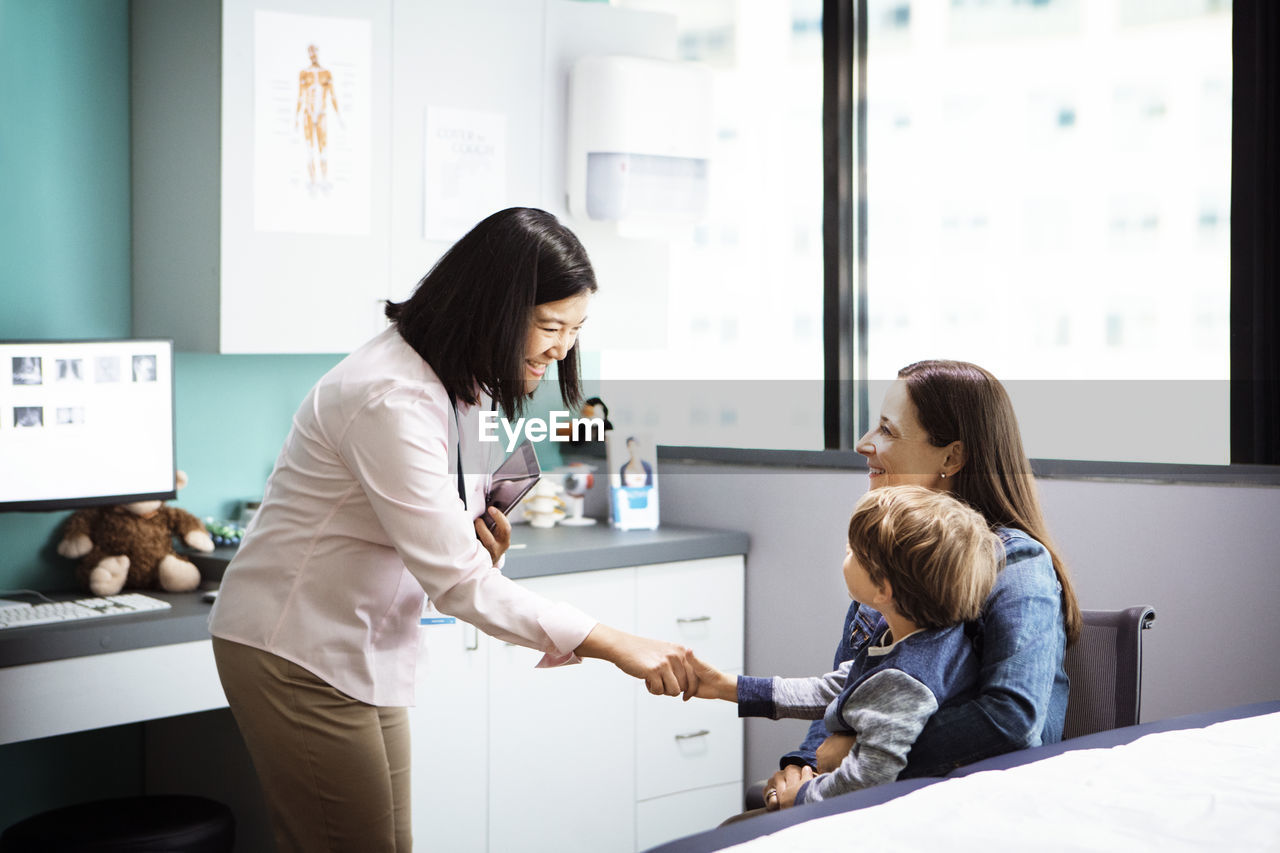 Happy female doctor greeting boy sitting with mother in clinic