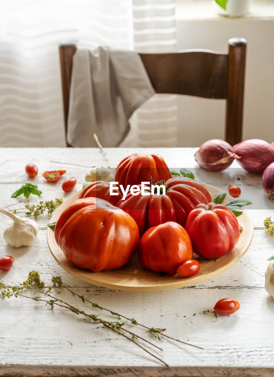 Red tomatoes on the plate and white wooden background with oregano, basil leaves, garlic and onion.