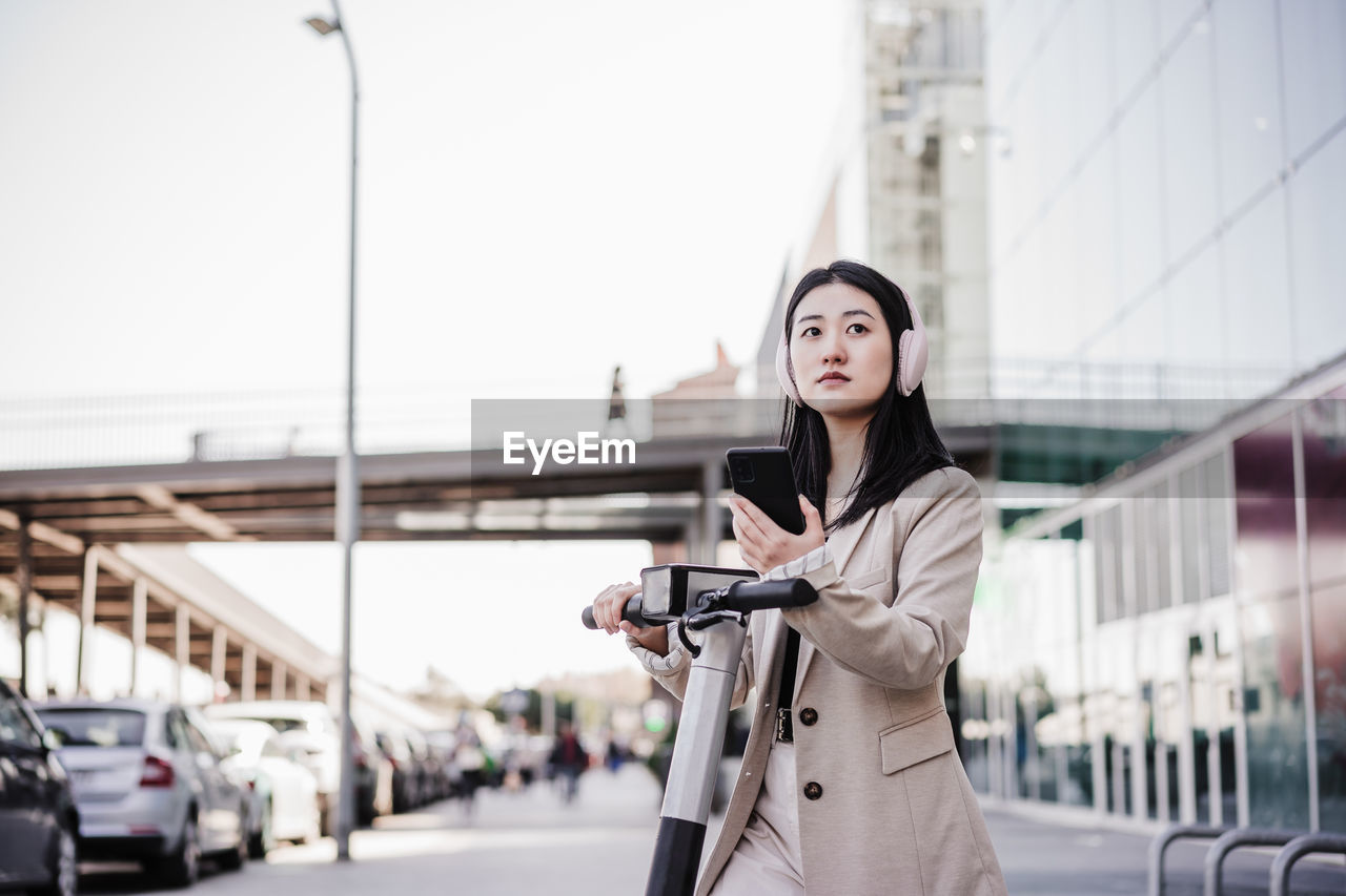 Chinese business woman renting electric scooter in city,wearing headphones, using mobile phone