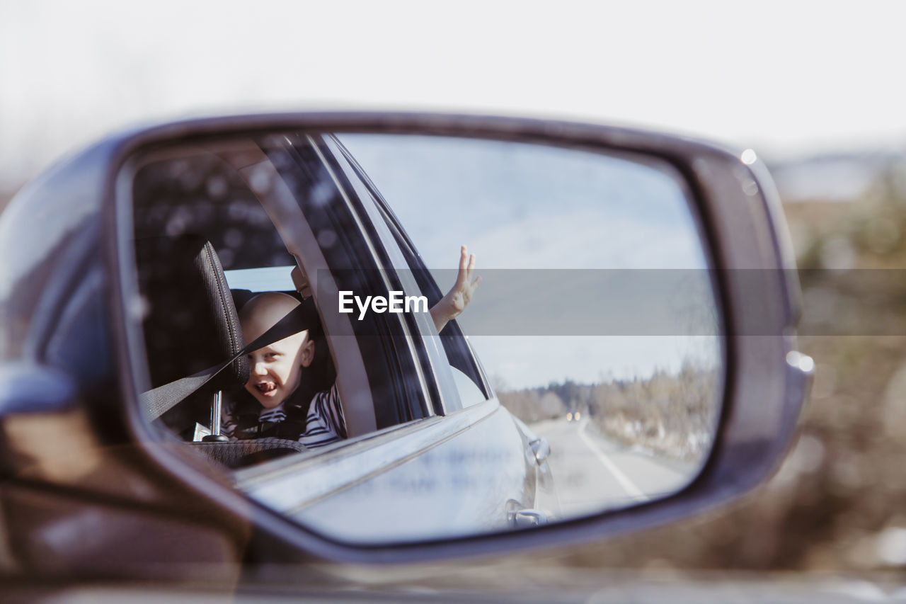 Car mirror shot of boy in back seat with hand out window on sunny day