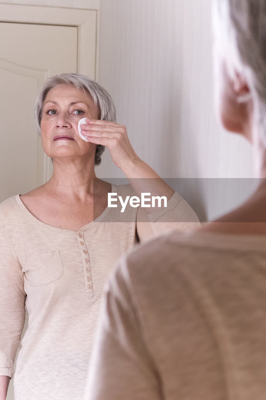 A elderly caucasian women with short gray hair cares for skin of face looking in the mirror at home