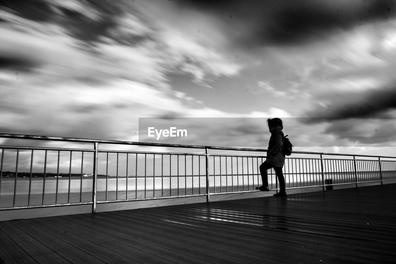 Full length of silhouette man standing on footpath while looking at sea against cloudy sky