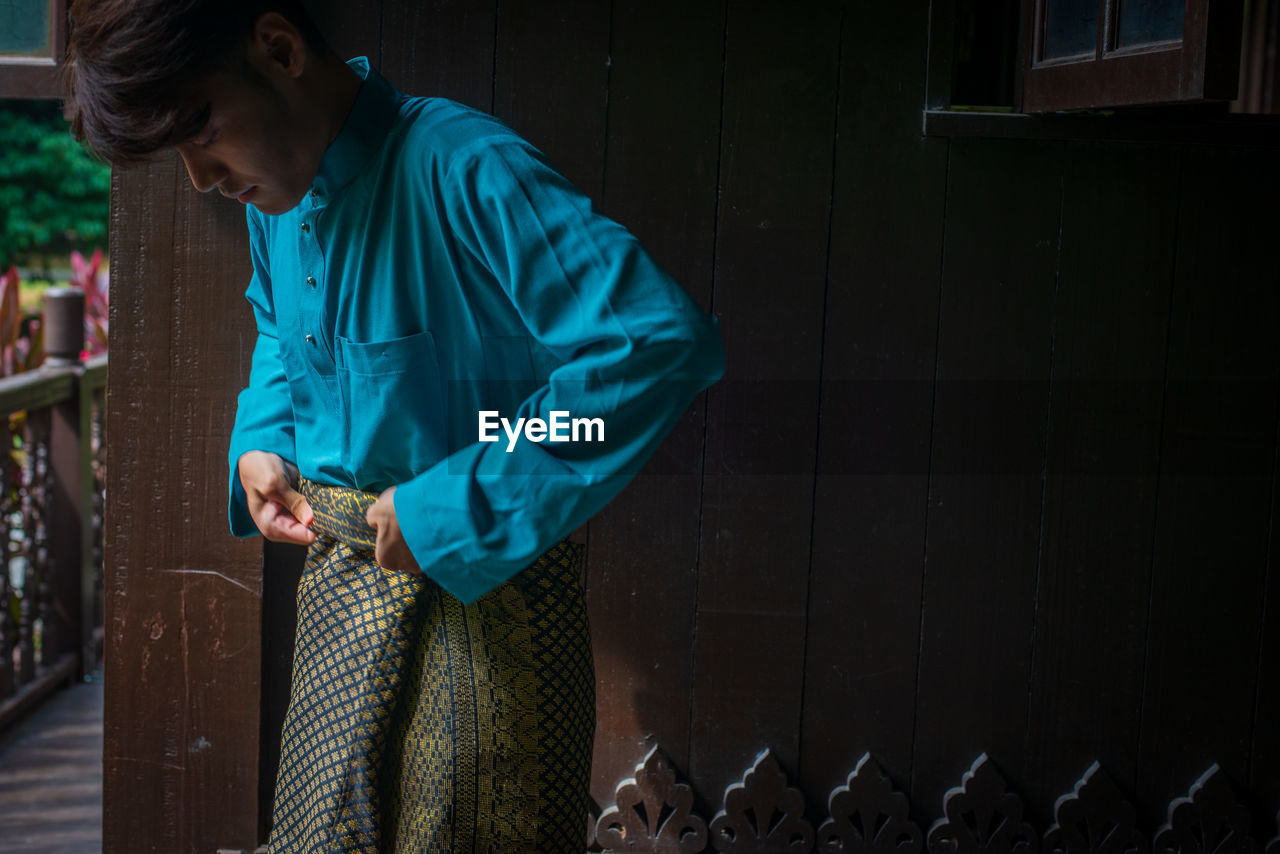 Side view of young man wearing sampin in traditional clothing in a malay wooden house during eid. 