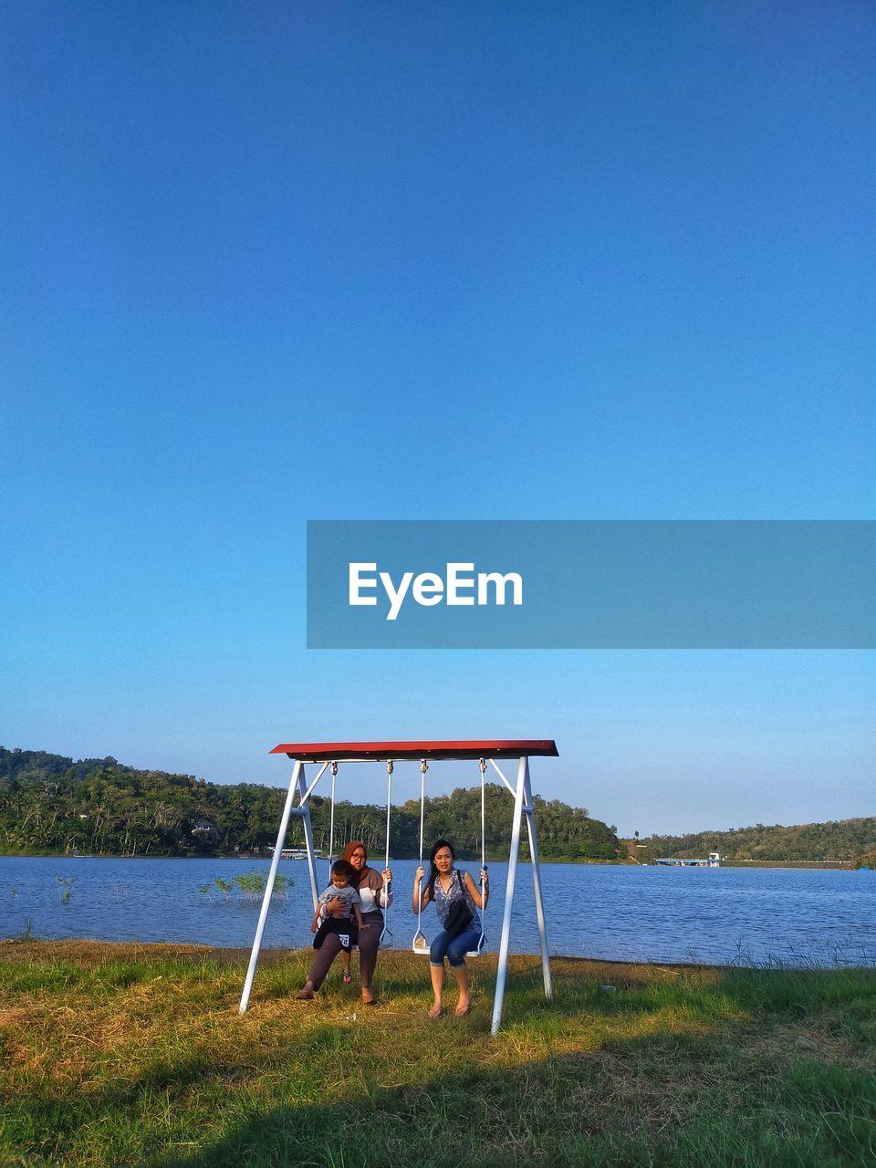 People sitting on grass at lake against clear blue sky