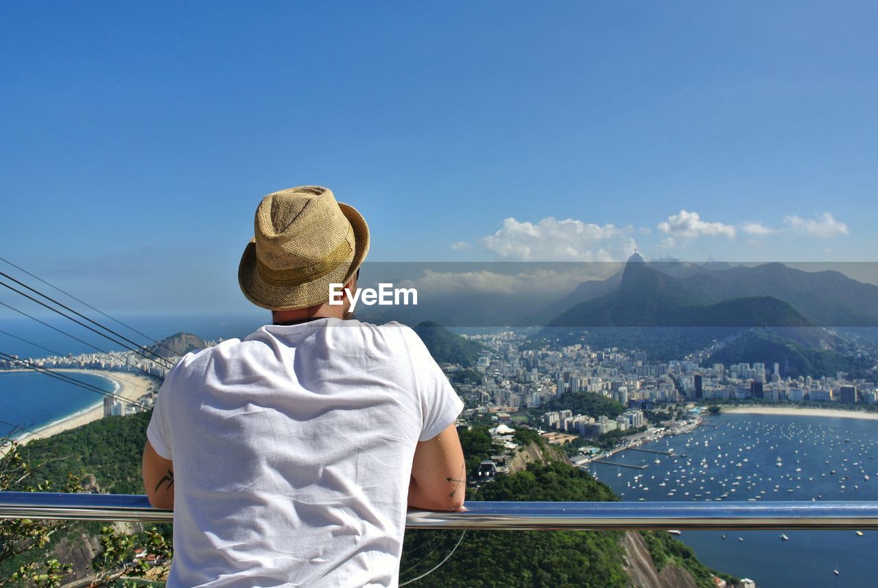 Rear view of man looking at cityscape while leaning on railing against sky