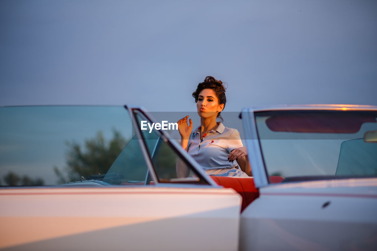 Portrait of young woman sitting on car against sky