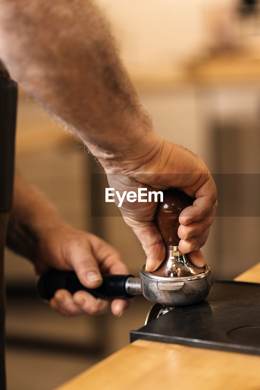 Side view of crop unrecognizable male barista pressing ground coffee with tamper in portafilter while standing at counter with mat during espresso preparation in cafe