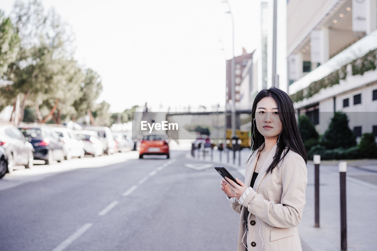 Confident business woman listening to music on headphones and mobile phone crossing street in city