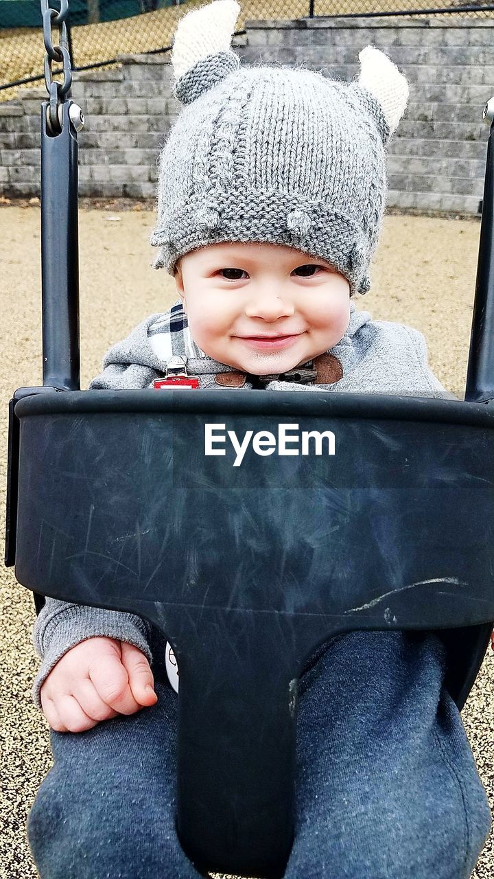 Portrait of cute smiling baby boy wearing warm clothing sitting on swing at playground