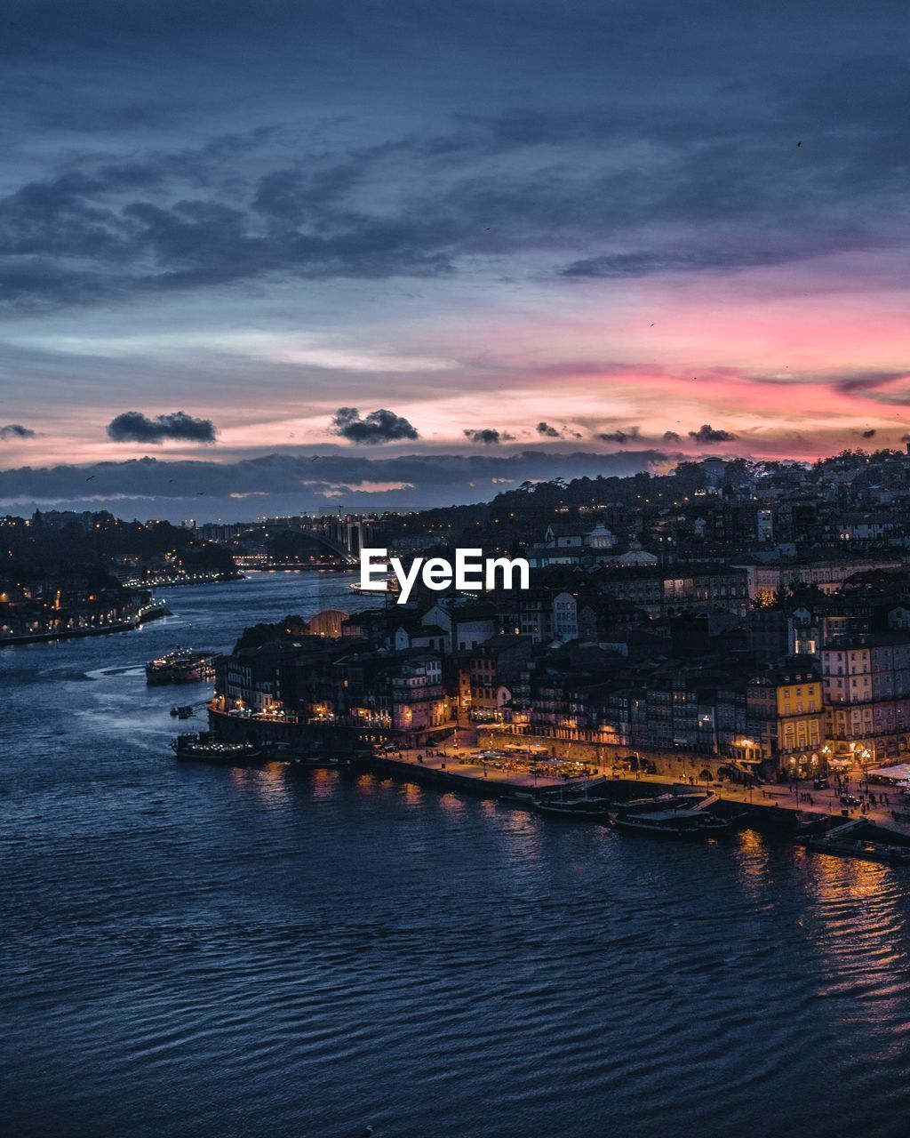 Illuminated city by sea against sky at sunset