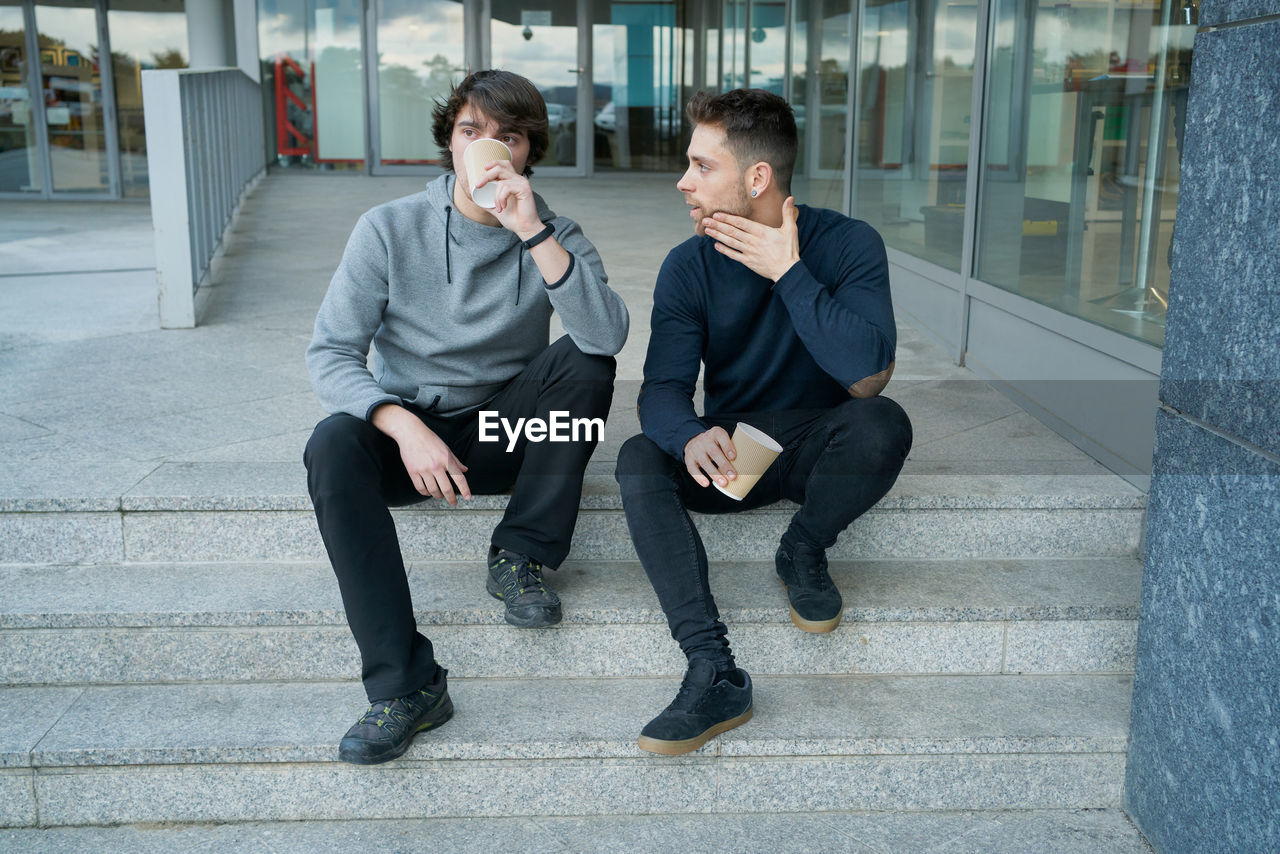 Front view of two young men talking sitting on a city staircase and lo