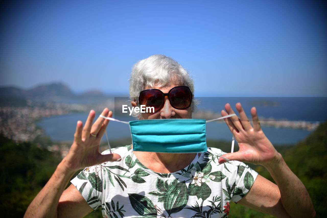 Portrait of elderly woman wearing sunglasses against sky showing mask