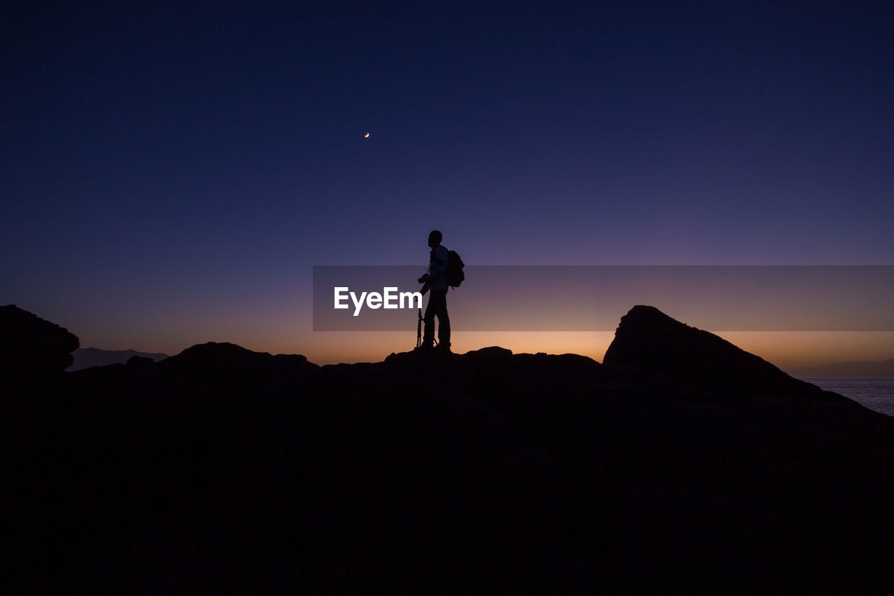 Silhouette photographer standing on rock against sky during sunset