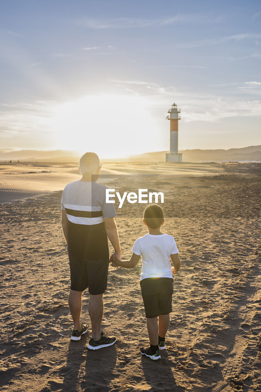 Rear view of man with son standing on beach against lighthouse